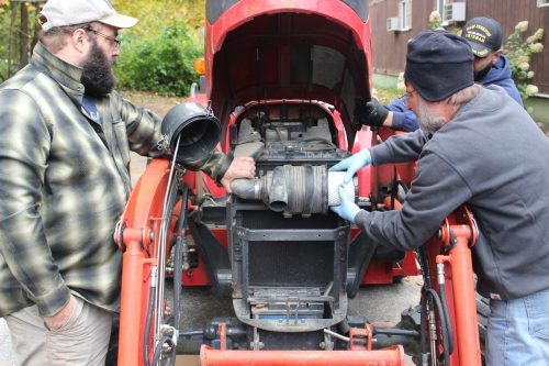 Two men on either side of a tractor with its hood open remove the air filter.